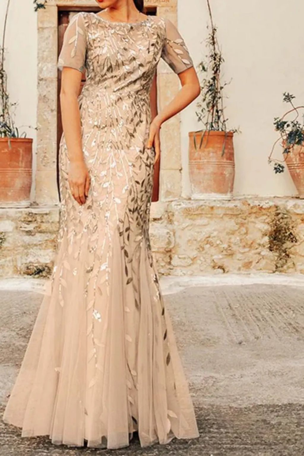 Full-length champagne trumpet formal dress displaying a deep V-neck, sheer long sleeves, and elegant gold-toned leaf embroidery trailing onto a flared tulle skirt