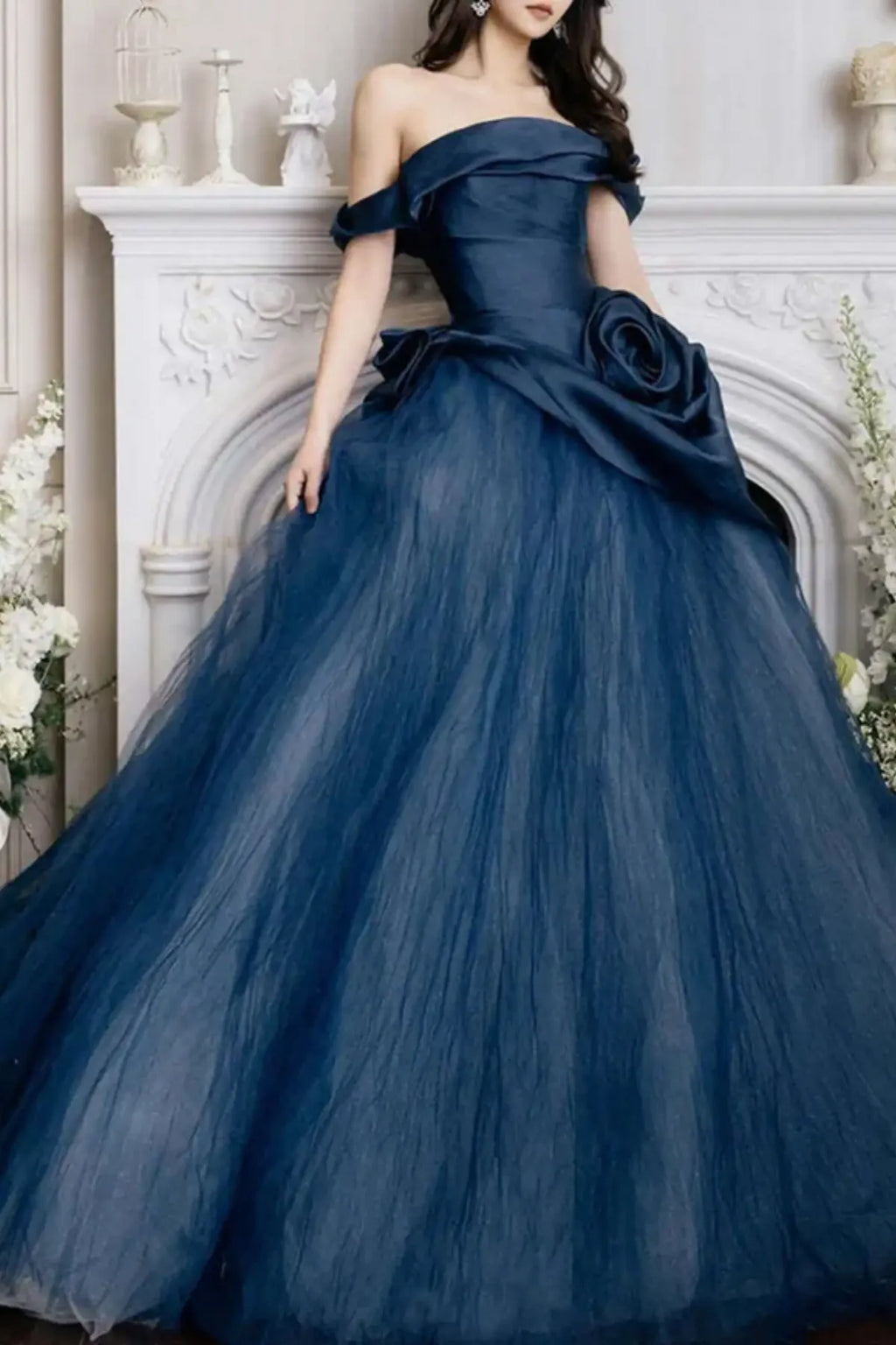 Full-length view of a model in a dramatic navy blue off-the-shoulder evening gown posing in front of an elegant white ornamental fireplace mantel