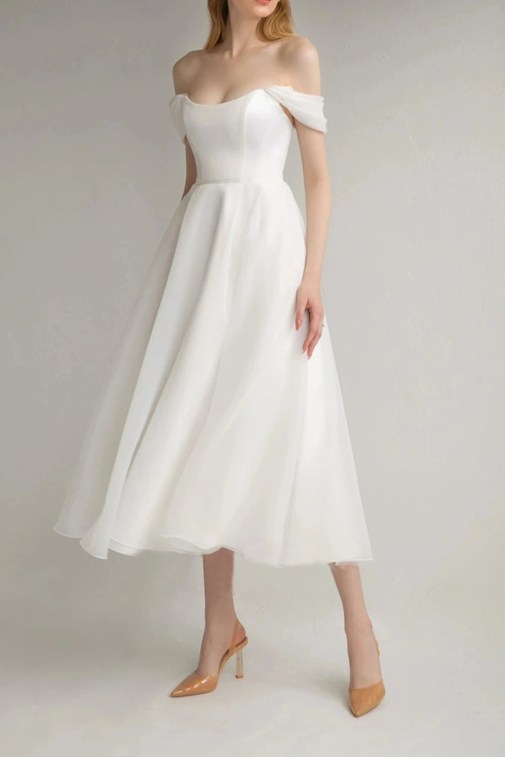 Full front view of a model wearing an ivory off-the-shoulder midi A-line bridal dress with nude heels
