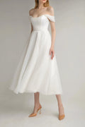 Full front view of a model wearing an ivory off-the-shoulder midi A-line bridal dress with nude heels