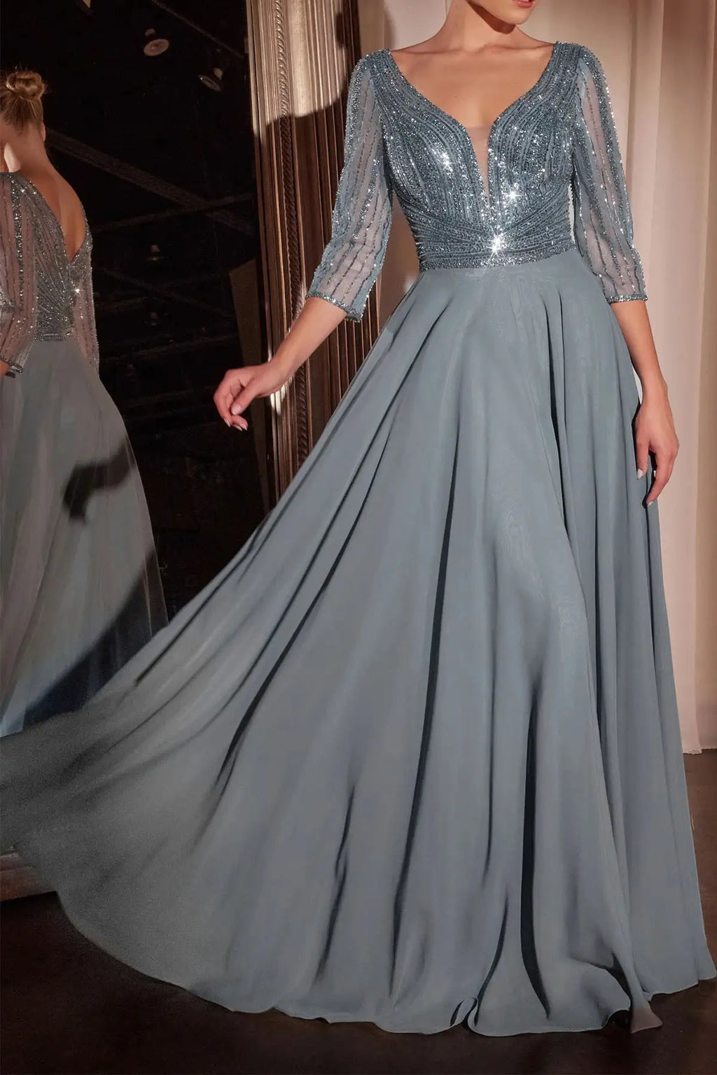A floor-length formal gown in dusty blue-gray with a heavily embellished sequined bodice, sheer three-quarter sleeves, and a full flowing skirt, shown on a model with a mirror reflection visible on the left.