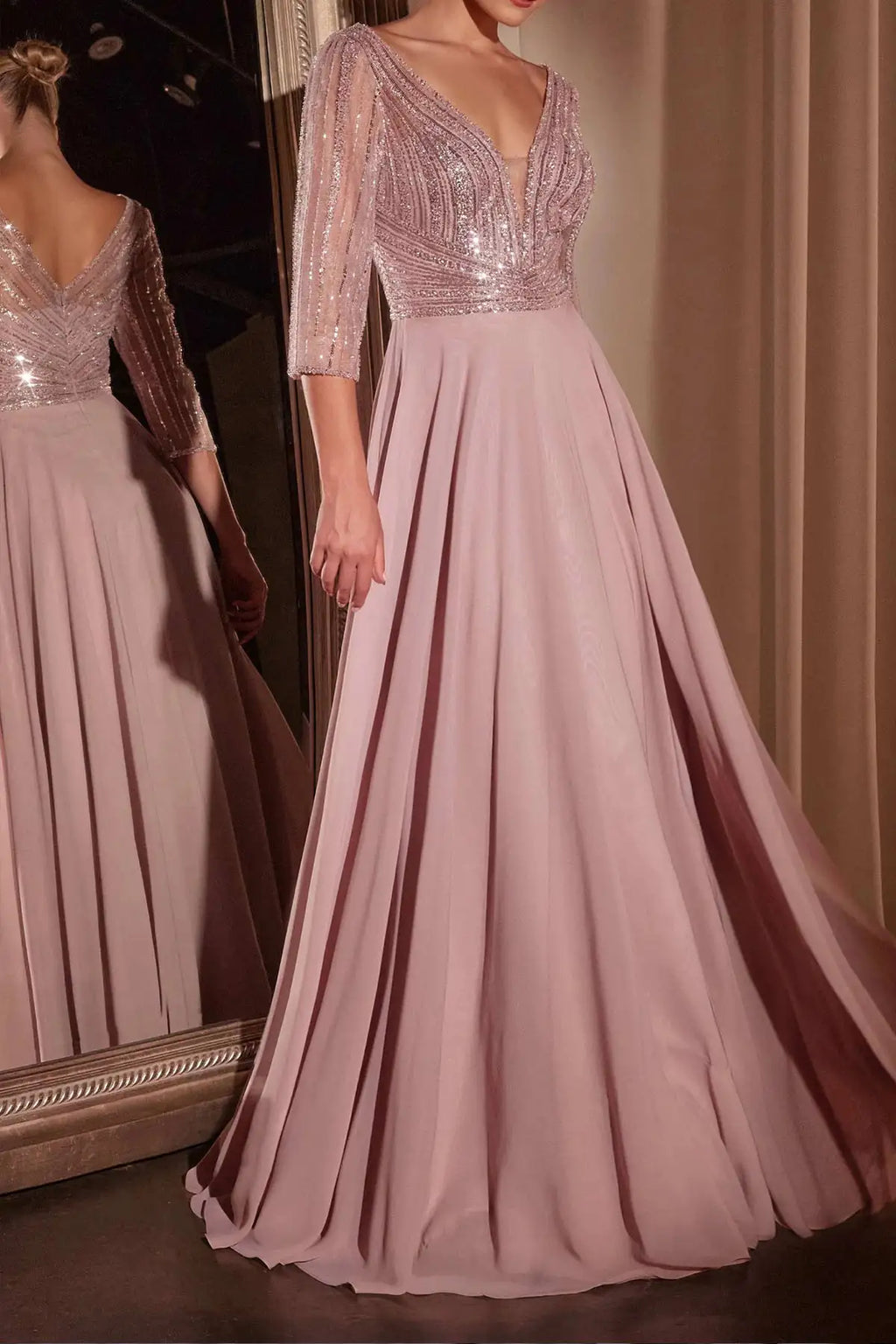A floor-length dusty pink evening gown with a deep V-neckline, embellished sequined bodice, and three-quarter sheer sleeves, shown on a model with a mirror reflection visible on the left.
