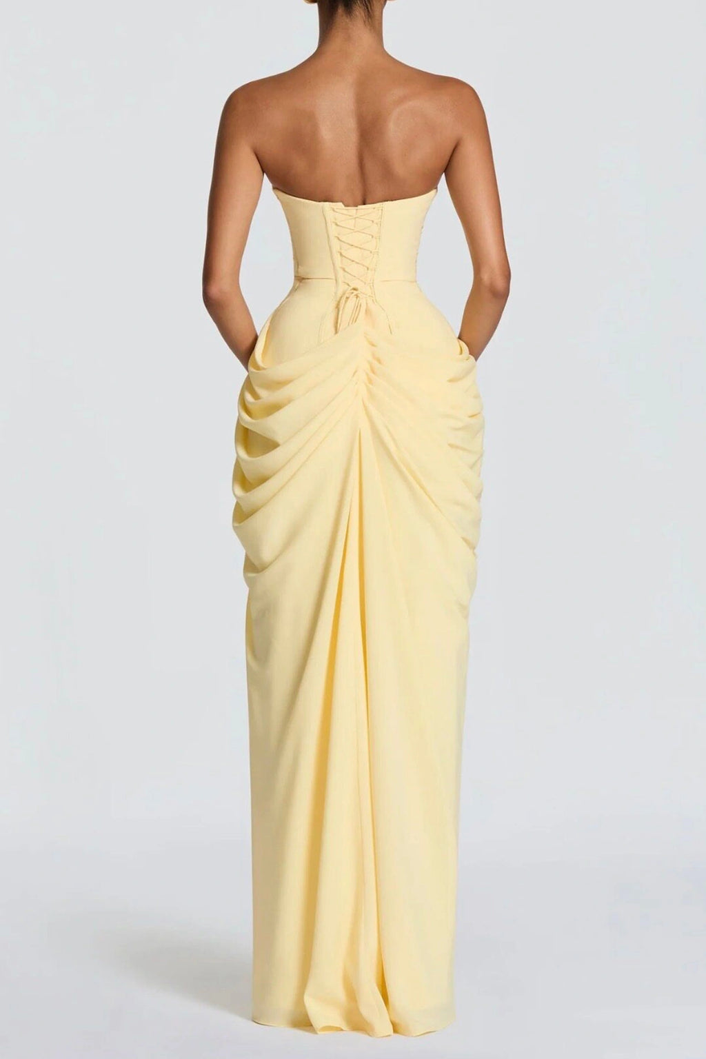 Studio back view of a pastel yellow strapless evening gown showcasing a structured lace-up corset and flowing rear drape